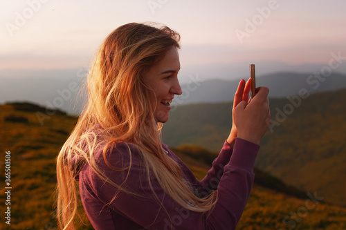 Young blonde woman making selfie on smartphone high in the mountains at sunset.