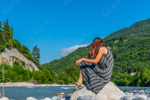 An unrecognizable relaxed young Caucasian redhead woman sitting on a boulder with a beer bottle next to the Var river in the French Alps enjoying summer sunshine