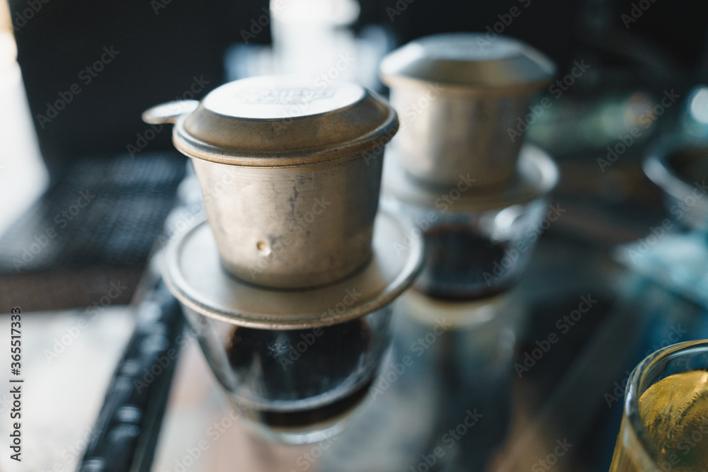 Hot milk coffee dripping in Vietnam style, pouring water over ground coffee contained in Vietnamese Phin Filter on table. Selective Focus. Hot milk coffee dripping in Vietnam style, pouring water over ground coffee contained in Vietnamese Phin Filter on table. Selective Focus.