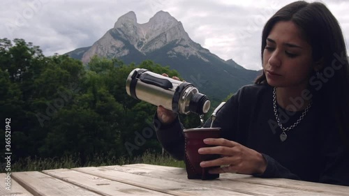Young woman preparing Argentinean Mate (Mate Argentino) outdoors in a wooden camping table. Girl pouring hot water in mate. Beautiful mountain landscape background
