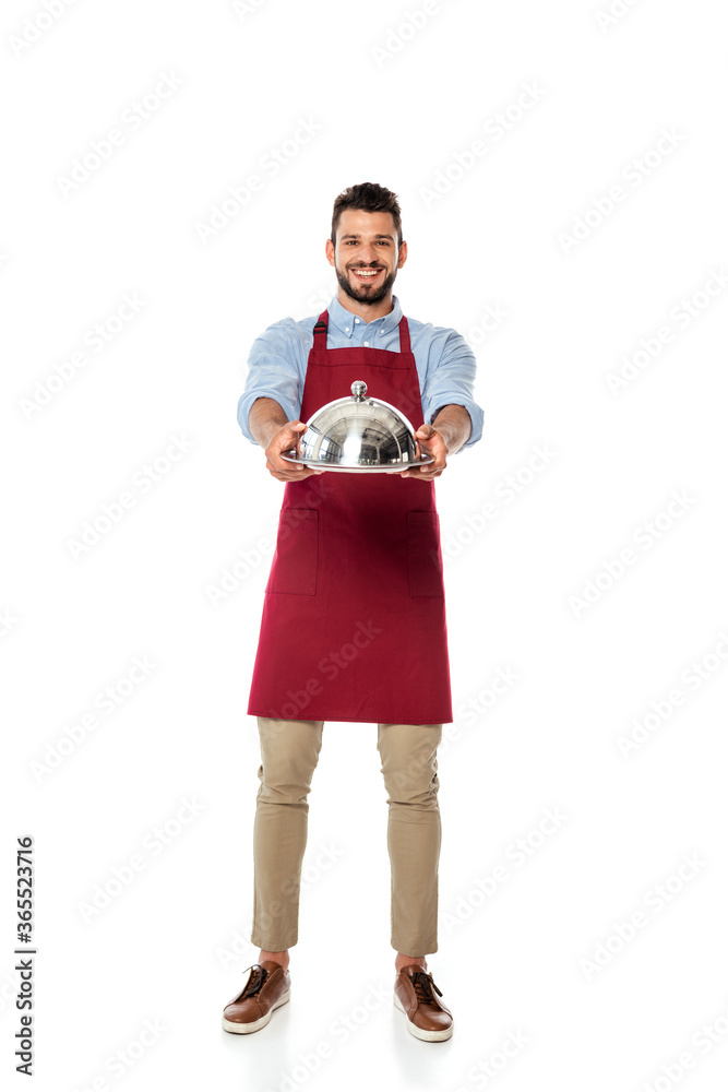 Handsome waiter smiling at camera and showing metal tray and dish cover on white background