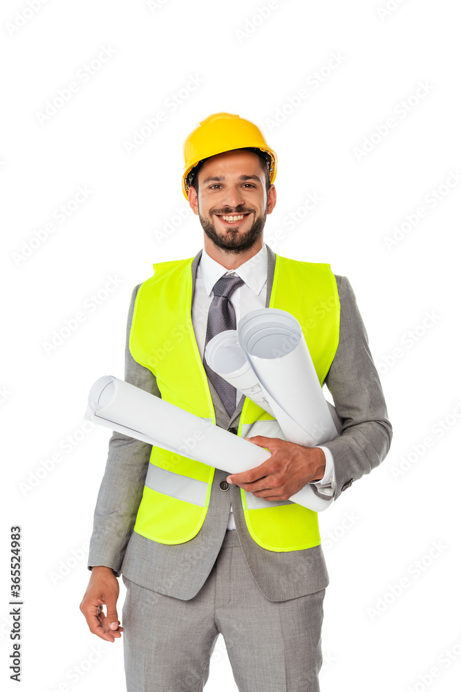 Handsome positive engineer in suit and safety vest holding blueprints and looking at camera isolated on white