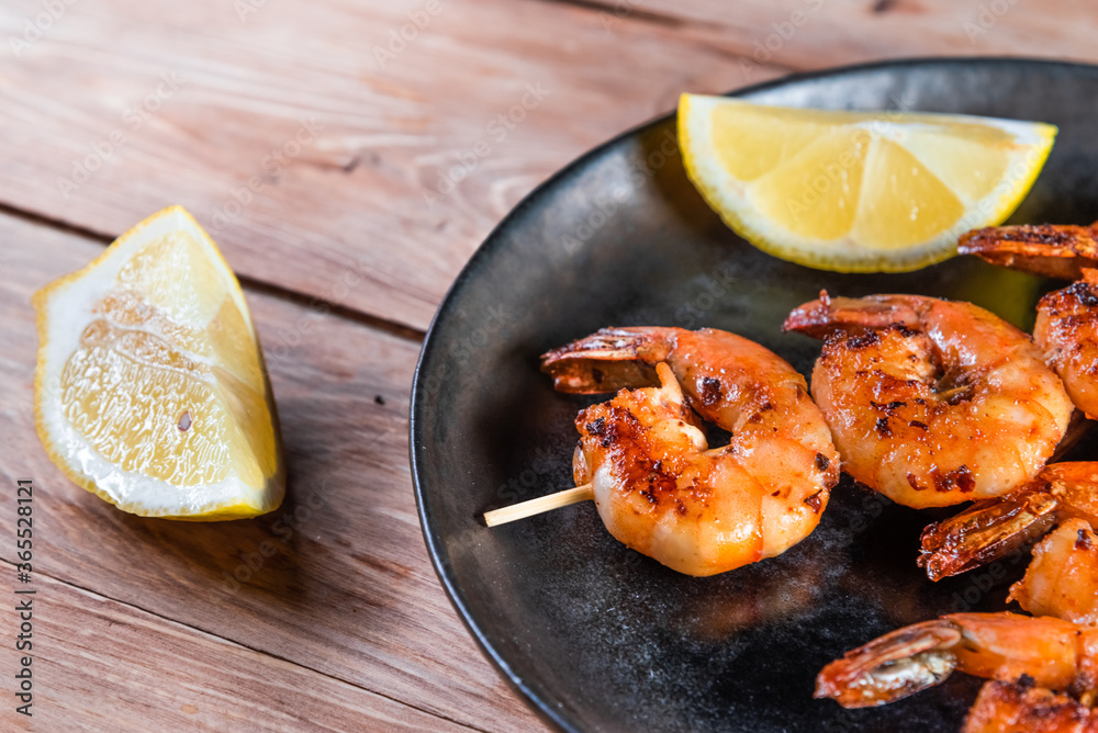 Appetizing fried shrimp on a skewer in a black plate on a wooden surface among lemon and seasonings. The concept of fast and wholesome food