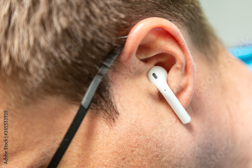 A wireless white small earpiece earphone in the ear of a man in black glasses close-up from the side. Horizontal orientation. 
