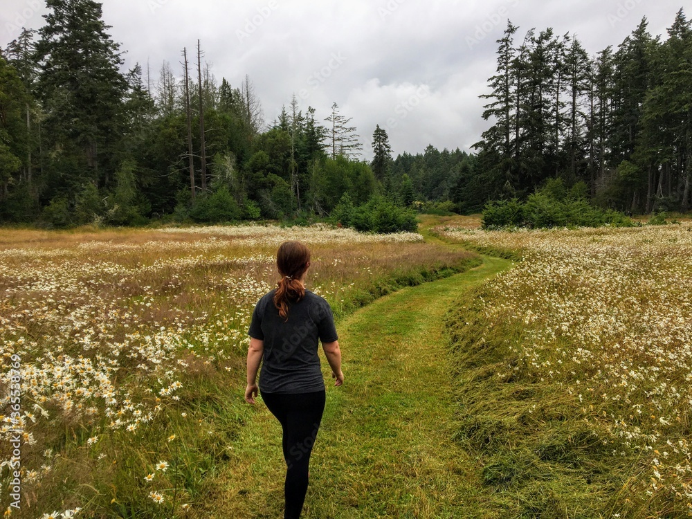 A young female hiker walking through the meadows of East Sooke Regional Park, surrounded by forest.  On Vancouver Island, British Columbia, Canada.