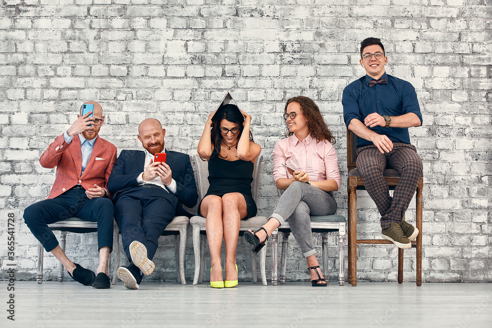 © Georgii - Group of smiling young business people sitting in line using mobile phones and other devices over white brick background © Georgii - Group of smiling young business people sitting in line using mobile phones and other devices over white brick background
