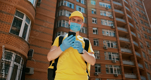 The delivery man in yellow uniform stands with a smartphone against the background of an apartment building, in medical gloves and a mask