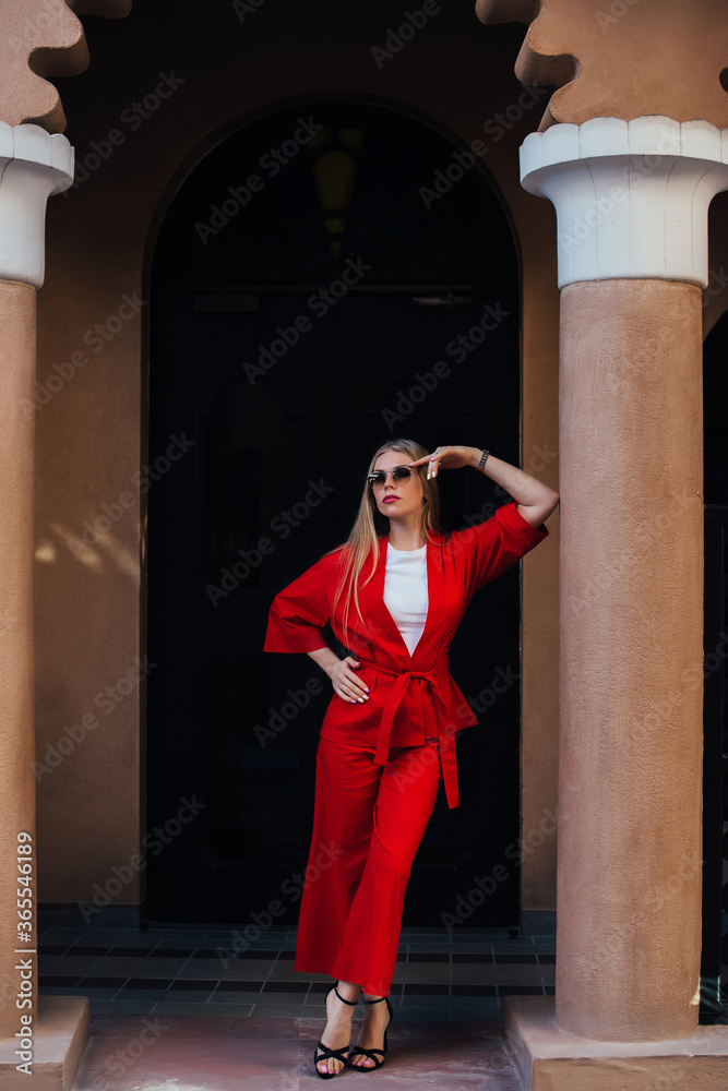 Portrait of a young blonde girl in a red suit who poses leaning on a column in the arch. A girl in red roll-top pants and a jacket, high-heeled shoes.