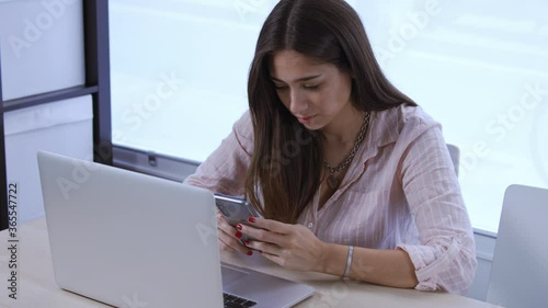 Beautiful young businesswoman wearing pink shirt working remotely from home using laptop pc and smartphone device. Female freelancer typing text message, checking social media or having video call
