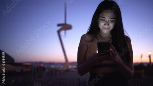 Smiling attractive young woman wearing sportswear, having break after workout or running, using Mobile Phone During Sunset. Sporty Girl typing a sms, texting friends, checking social media

