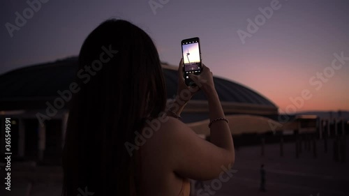 Attractive young woman takes picture with smartphone device to social account or video stories. Beautiful girl taking photo with mobile phone at sunset wearing sportswear having break after running
