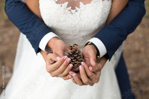 Groom embraces bride in a pine forest, their hands holding a lump