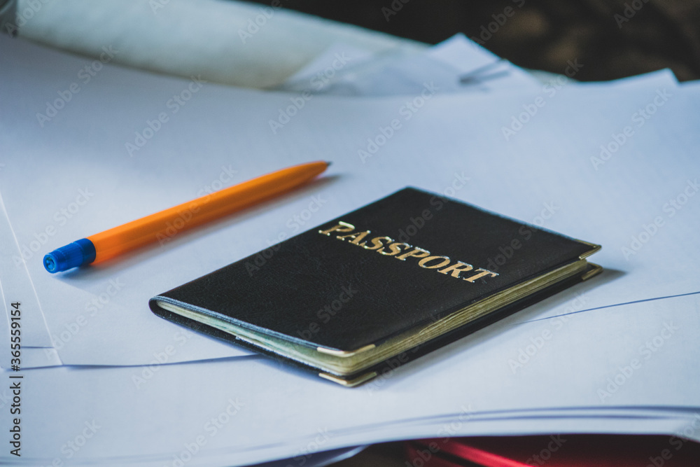 A passport with a black cover lies on white documents next to a ballpoint pen. A passport with a black cover lies on white documents next to a ballpoint pen.
