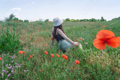 Young unknowable girl in hat sitting back to us in a field with poppies and and dreams, sunny day, rural lifestyle in summer