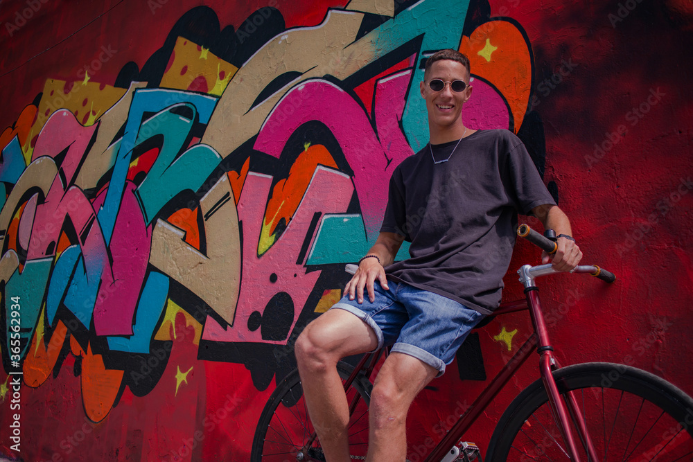 Obraz premium a guy with round sunglasses in a dark t-shirt and shorts with a bicycle against a bright wall with graffiti