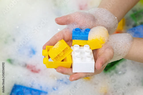 Toys washing. Child hands with colorful bright construction blocks in soap foam water. Cleaning children toys idea. Corona virus COVID-19 protection concept.