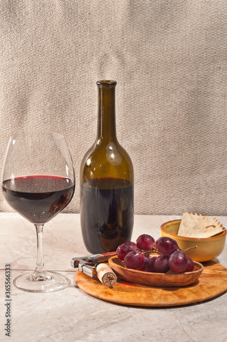 front view, medium distance of a glass of red wine, wine bottle, rare wood bowl of freshly picked, washed red grapes, cork, corkscrew and a bowl of crackers, for a wine tasting event