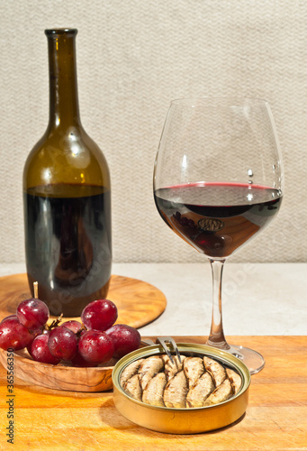 front view, medium distance of a glass of red wine, wine bottle, rare wood plate, freshly picked, washed red grapes in rare wood bowl and a can of sardienes, for a wine tasting event