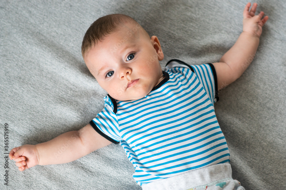 Cute caucasian, blue eyed baby boy lying down on gray blanket with his arms spread open. Baby in striped outfit.