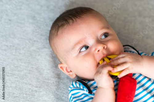 Obraz na płótnie cute caucasian baby boy with blue eyes in striped clothes biting a toy teether, lying down on gray blanket. Teething solution. w salonie