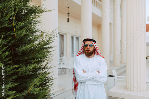 Handsome middle eastern arab muslim  man in traditional clothing standing in front of the white beautiful modern house photo