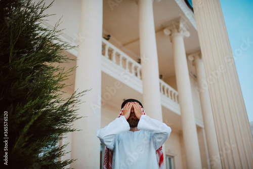 Handsome middle eastern arab muslim man in traditional clothing standing in front of the white beautiful modern house with his hands over his face