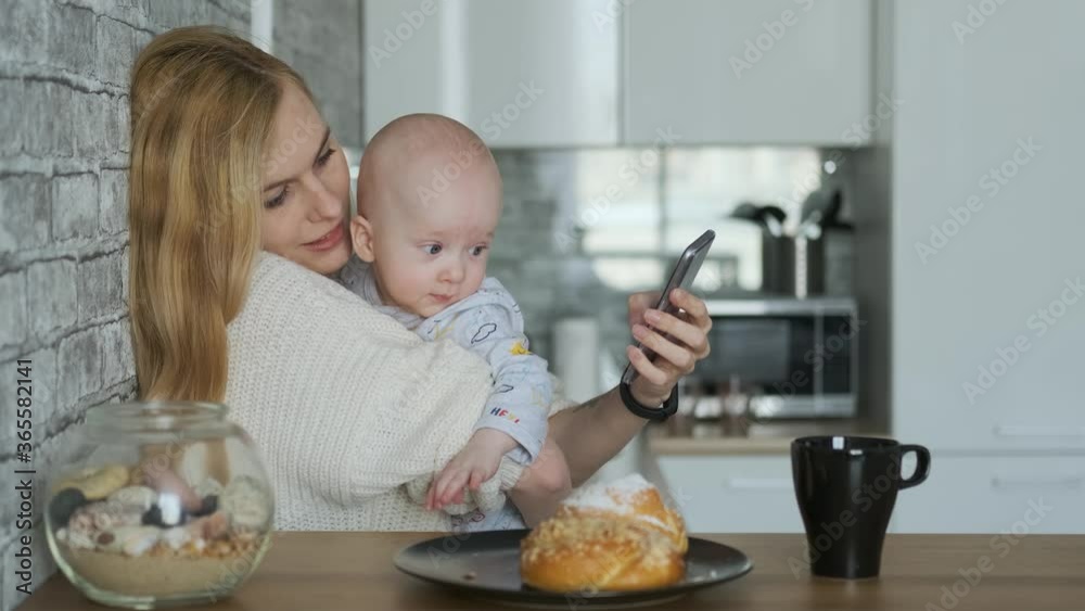 mom with baby in her arms uses a smartphone while sitting in the kitchen mom with baby in her arms uses a smartphone while sitting in the kitchen