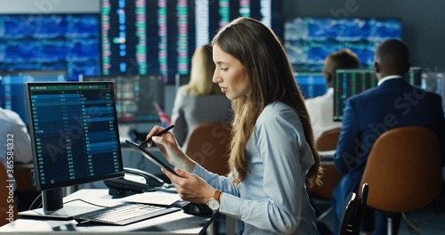 Tableau sur toile Female stock trader working at stock exchange office using digital tablet computer on background of multiple monitors showing data, ticker numbers and graphs