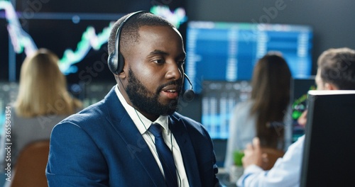 Portrait of african american trader or broker working at stock exchange office using headset and computer on background of his business team. Investment Entrepreneur Trading Concept.