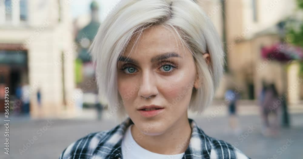 Portrait of young Caucasian blonde girl with nose piercing standing in beautiful city outdoors looking at camera with smile. Close up of pretty woman in hipster style smiling. Female face zooming in. Portrait of young Caucasian blonde girl with nose piercing standing in beautiful city outdoors looking at camera with smile. Close up of pretty woman in hipster style smiling. Female face zooming in.