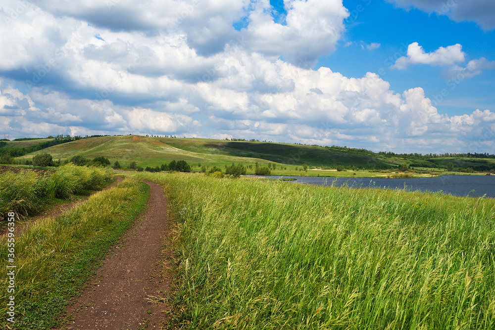 A country road among fields with grasses, near a lake and hills. Summer natural landscape with cloudy sky. A country road among fields with grasses, near a lake and hills. Summer natural landscape with cloudy sky.