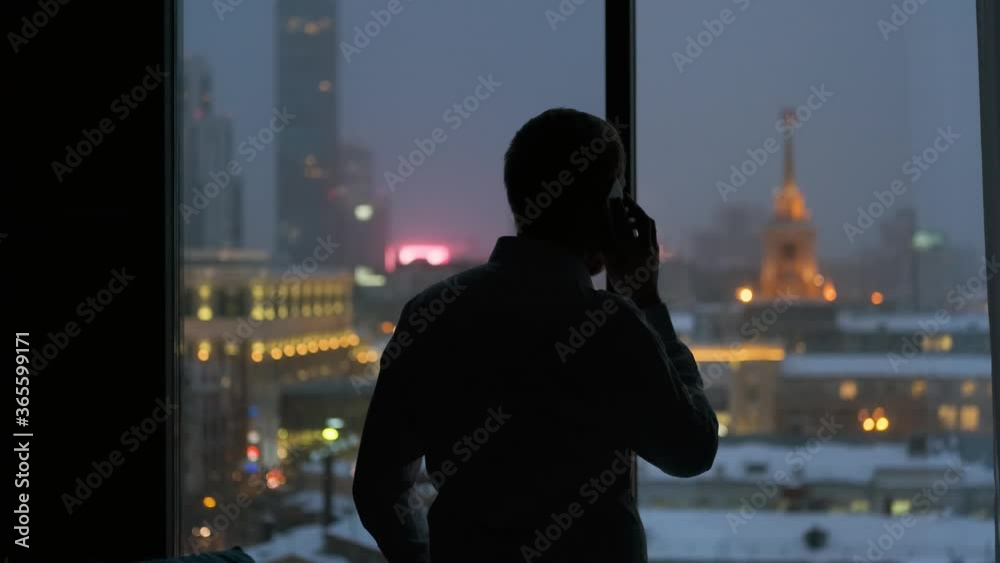 businessman talking on the phone by the window overlooking the night city businessman talking on the phone by the window overlooking the night city