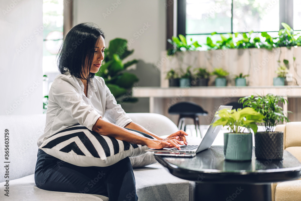 Smiling beautiful balck business woman wearing white shirt relaxing using laptop.Young hipster girl freelancer working and sitting on sofa in green plant terrace decorated office