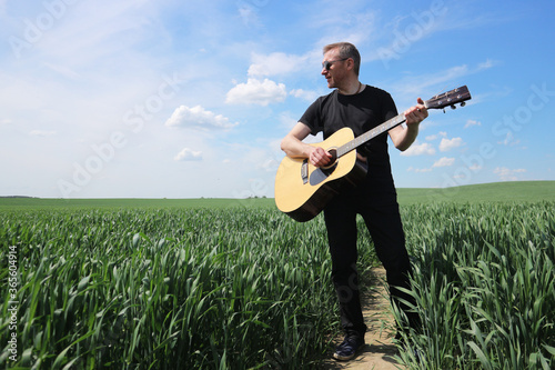 A man in black sunglasses with a guitar in the field. One man sings in nature against a blue sky. Romantic guitarist plays and goes along the path.