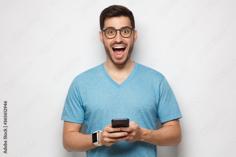 Young handsome manin blue t-shirt, wearing eyeglasses, holding phone in hands, looking at camera with excited WOW face, isolated on gray background Young handsome manin blue t-shirt, wearing eyeglasses, holding phone in hands, looking at camera with excited WOW face, isolated on gray background