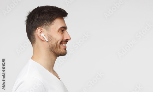 Horizontal banner of young smiling man in white t-shirt, enjoying music through wireless earphones, isolated on gray background with copy space