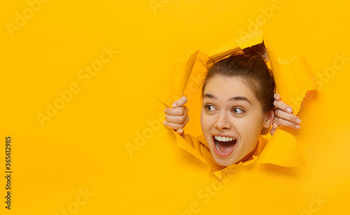 Excited girl looking through hole in paper at left with eyes round with surprise, isolated on yellow background with copy space photo