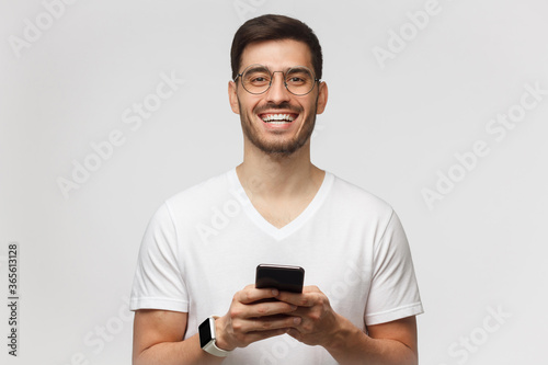 Portrait of handsome young man in white t-shirt, holding smartphone, looking at camera and laughing, isolated on gray background © Damir Khabirov