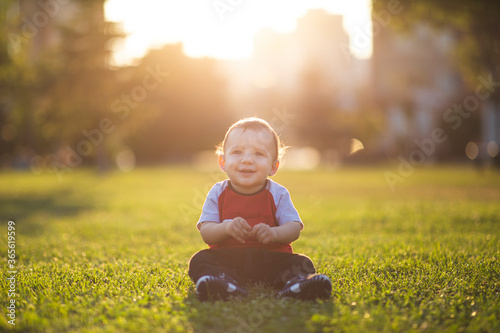 a portrait of a smiling baby, he is sitting on a green grass and thinks in a positive mood.