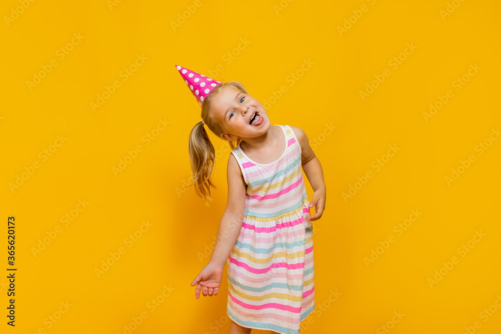 Happy birthday child girl with two ponytales in pink cap on colored yellow background showing her tongue.