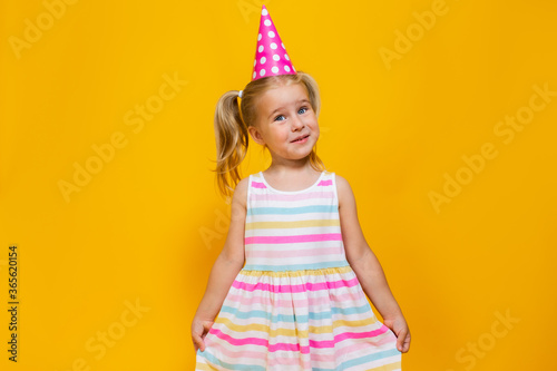 Happy birthday child girl with two ponytales in pink cap holding her dress on colored yellow background.