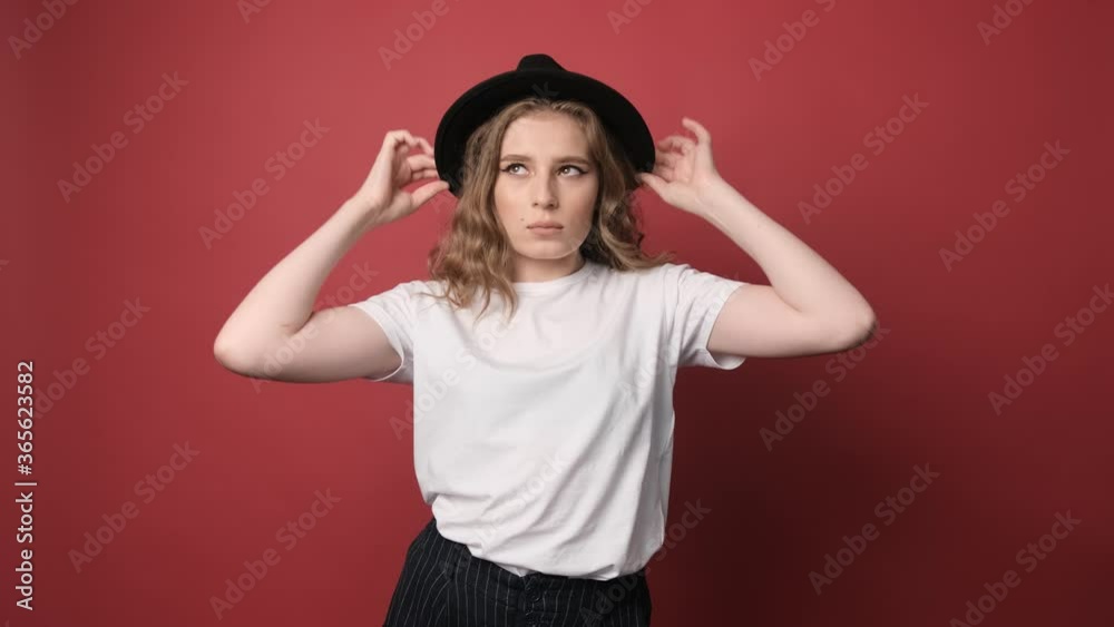 Attractive young woman with a curly hair, dressed in a white t shirt is playing with a black hat over red background Attractive young woman with a curly hair, dressed in a white t shirt is playing with a black hat over red background