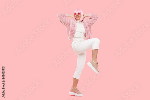Full length portrait of young happy girl dancing, wearing funky clothes and glasses, lifting knees and jumping, isolated on pink background