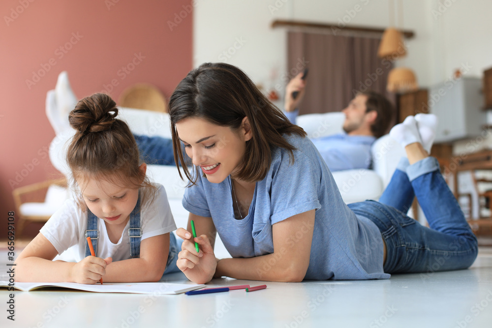 Happy mother smilling daughter lying on warm floor enjoying creative activity, drawing pencils coloring pictures in albums, father resting on couch, family spend free time together. Happy mother smilling daughter lying on warm floor enjoying creative activity, drawing pencils coloring pictures in albums, father resting on couch, family spend free time together.