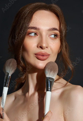 Portrait of the beautiful brunette woman with make-up brushes near face. Girl posing over Gray background. Clean natural makeup.