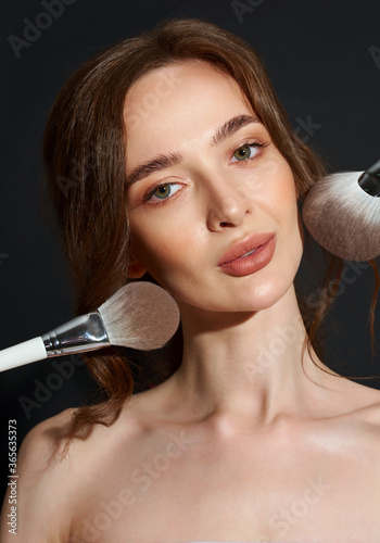Portrait of the beautiful brunette woman with make-up brushes near face. Girl posing over Gray background. Clean natural makeup.