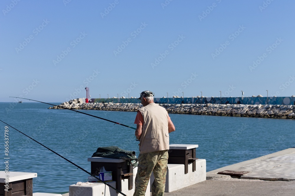 Obraz premium Pesaro, Italy - 9 july 2020: an old fisherman in fishing on the pier of the harbor