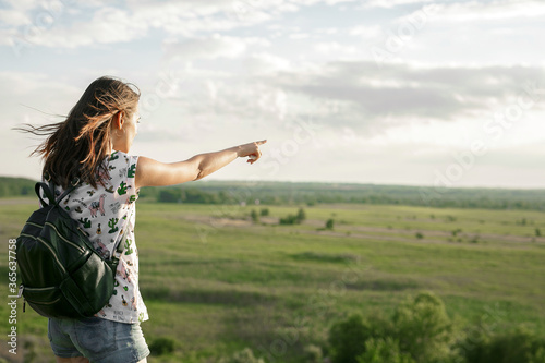 Young Happy Woman Traveler with backpack showing something far beyond the horizon and relaxing outdoor with rocky mountains on green field background. Summer vacations and Lifestyle hiking concept.