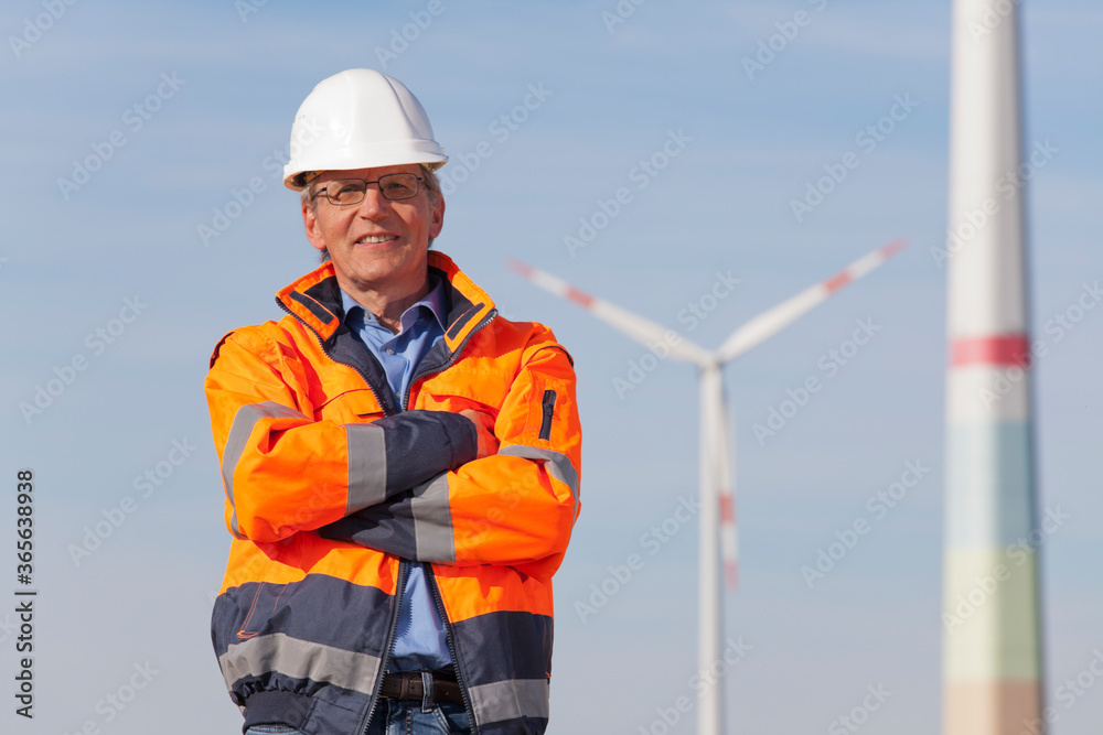 Smiling engineer with hard hat and protective clothing in front of wind turbines on a sunny day Smiling engineer with hard hat and protective clothing in front of wind turbines on a sunny day