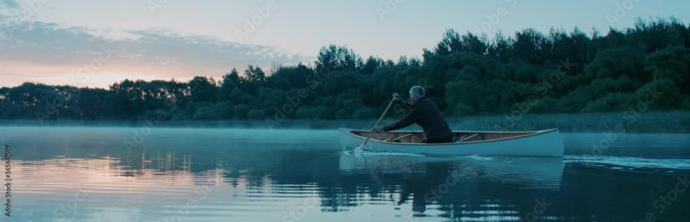 Man canoeing in a traditional wooden boat on a large lake at dawn. Shot on RED cinema camera with 2x anamorphic lens Man canoeing in a traditional wooden boat on a large lake at dawn. Shot on RED cinema camera with 2x anamorphic lens